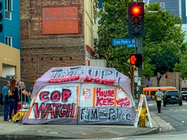 A street corner with an urban setting, featuring a large tent with bold graffiti-style writing advocating social issues. Behind the tent is a brick building and a traffic light showing red. There are several people standing nearby, and a bus is visible in the background.