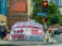 A street corner with an urban setting, featuring a large tent with bold graffiti-style writing advocating social issues. Behind the tent is a brick building and a traffic light showing red. There are several people standing nearby, and a bus is visible in the background.