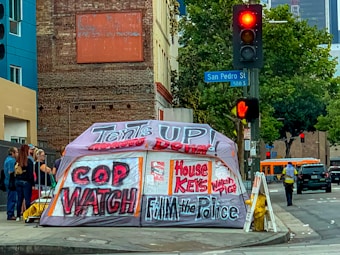 A street corner with an urban setting, featuring a large tent with bold graffiti-style writing advocating social issues. Behind the tent is a brick building and a traffic light showing red. There are several people standing nearby, and a bus is visible in the background.