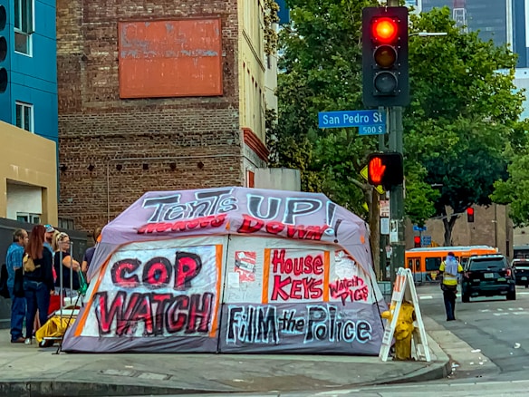 A street corner with an urban setting, featuring a large tent with bold graffiti-style writing advocating social issues. Behind the tent is a brick building and a traffic light showing red. There are several people standing nearby, and a bus is visible in the background.