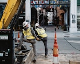 Two construction workers wearing safety vests and helmets are working on a street. They are standing beside a construction vehicle, surrounded by orange traffic cones. The background shows a narrow alleyway with a colorful, geometric arch overhead. Buildings line both sides of the alley, and string lights are hanging above the pathway.