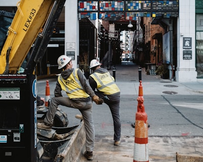 Two construction workers wearing safety vests and helmets are working on a street. They are standing beside a construction vehicle, surrounded by orange traffic cones. The background shows a narrow alleyway with a colorful, geometric arch overhead. Buildings line both sides of the alley, and string lights are hanging above the pathway.
