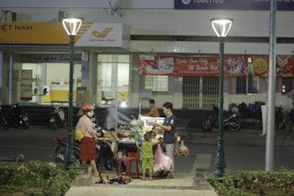 A street food vendor serves two customers, a woman and a child, at a cart under streetlights. The scene is in front of a Vietnam Post office with motorcycles parked nearby. A red banner is displayed on the building behind them.