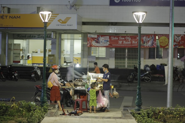 A street food vendor serves two customers, a woman and a child, at a cart under streetlights. The scene is in front of a Vietnam Post office with motorcycles parked nearby. A red banner is displayed on the building behind them.
