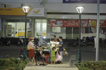 A street food vendor serves two customers, a woman and a child, at a cart under streetlights. The scene is in front of a Vietnam Post office with motorcycles parked nearby. A red banner is displayed on the building behind them.