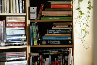 A well-organized bookshelf filled with a variety of books, including art books, mythology, and history. The books are aligned on wooden shelves, with some stacked horizontally. A green vine plant decorates the right side of the shelf, adding a natural element to the setting.