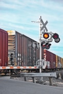 A freight train moves past a railroad crossing with red warning lights activated. The scene includes a metal crossing sign and barrier arm, set against a clear blue sky.