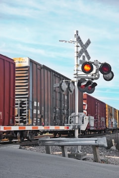 A freight train moves past a railroad crossing with red warning lights activated. The scene includes a metal crossing sign and barrier arm, set against a clear blue sky.