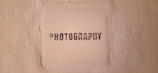 The image features a textured beige wall with a square area in the center that has the word 'PHOTOGRAPHY' stenciled in black, capital letters.