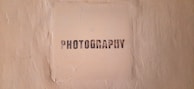 The image features a textured beige wall with a square area in the center that has the word 'PHOTOGRAPHY' stenciled in black, capital letters.