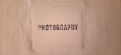 The image features a textured beige wall with a square area in the center that has the word 'PHOTOGRAPHY' stenciled in black, capital letters.