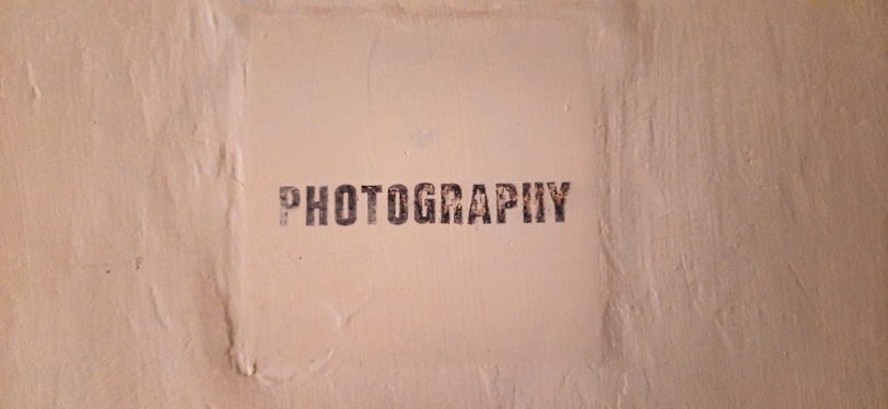 The image features a textured beige wall with a square area in the center that has the word 'PHOTOGRAPHY' stenciled in black, capital letters.