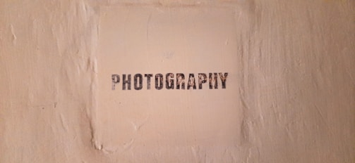 The image features a textured beige wall with a square area in the center that has the word 'PHOTOGRAPHY' stenciled in black, capital letters.