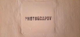 The image features a textured beige wall with a square area in the center that has the word 'PHOTOGRAPHY' stenciled in black, capital letters.