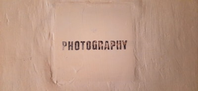 The image features a textured beige wall with a square area in the center that has the word 'PHOTOGRAPHY' stenciled in black, capital letters.