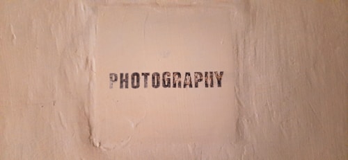 The image features a textured beige wall with a square area in the center that has the word 'PHOTOGRAPHY' stenciled in black, capital letters.