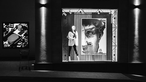 The image depicts a storefront display at night featuring a mannequin dressed in a suit positioned in front of a large art piece resembling a classical statue's face. The display window is bordered by geometric patterns and lit from above, creating dramatic shadows. To the left, a shopping cart is visible in front of a textured column, and another abstract art piece hangs on the wall.