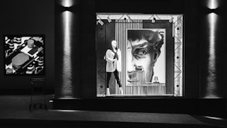 The image depicts a storefront display at night featuring a mannequin dressed in a suit positioned in front of a large art piece resembling a classical statue's face. The display window is bordered by geometric patterns and lit from above, creating dramatic shadows. To the left, a shopping cart is visible in front of a textured column, and another abstract art piece hangs on the wall.