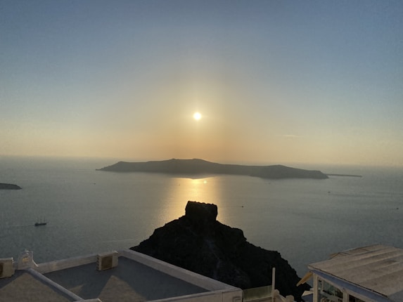 A serene sunset view over a calm sea with an island silhouette on the horizon. The sunlight creates a golden reflection on the water, and a few small boats are visible in the distance. In the foreground, there are rooftops with white surfaces typical of Mediterranean architecture.