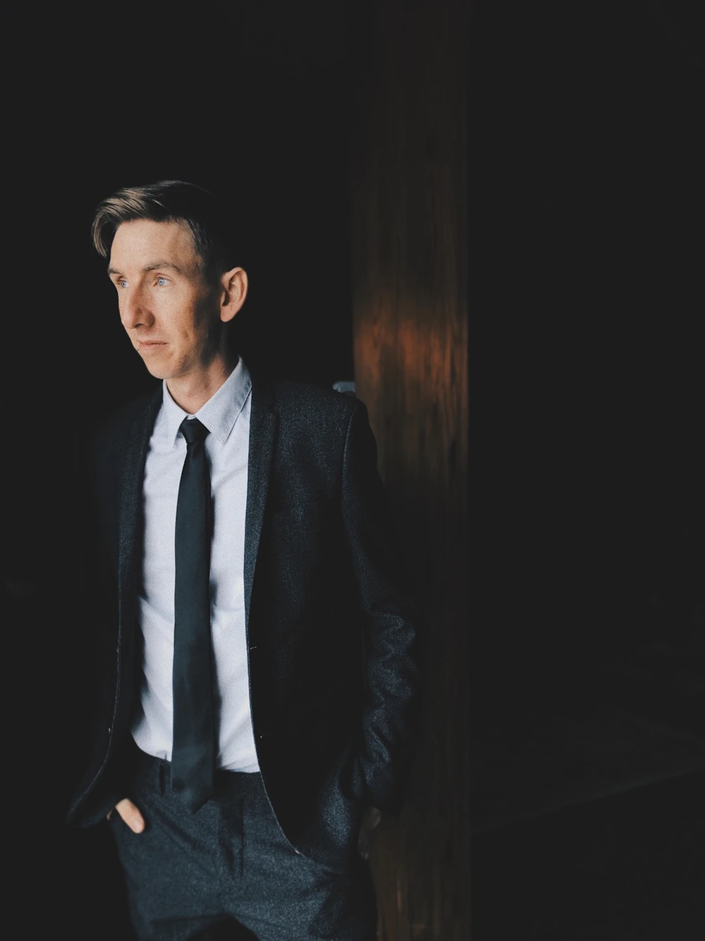 Editorial portrait of a man in a dark suit standing in low, dramatic light