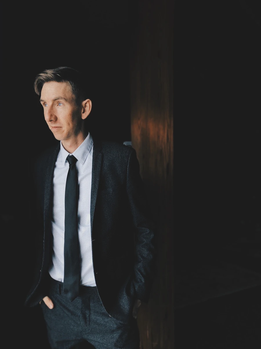 Editorial portrait of a man in a dark suit standing in low, dramatic light
