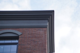 A corner view of a building featuring a combination of brick and concrete materials. The architecture includes a layered design at the roofline, with a large arched window visible. The sky is partly cloudy, providing a soft light to the structure.