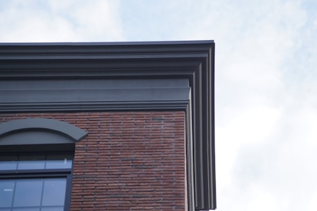 A corner view of a building featuring a combination of brick and concrete materials. The architecture includes a layered design at the roofline, with a large arched window visible. The sky is partly cloudy, providing a soft light to the structure.