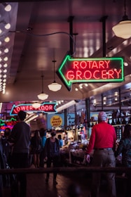 A bustling indoor market with neon signs indicating 'Rotary Grocery' and 'Lower Floor'. The environment is lively with people walking and shopping. The market is well-lit with hanging lights and displays signs of various stalls. Some shoppers are wearing masks, indicating a modern-day setting.