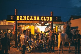 A lively scene in front of a venue called Bandana Club, featuring several people gathered outside. Some are standing near motorcycles, and a person on horseback is also present. The surroundings are illuminated with a warm, inviting glow coming from the club's lights. The ambiance suggests a festive or social gathering during nighttime.