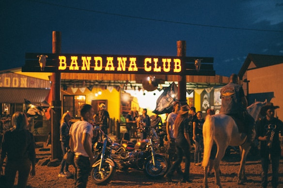 A lively scene in front of a venue called Bandana Club, featuring several people gathered outside. Some are standing near motorcycles, and a person on horseback is also present. The surroundings are illuminated with a warm, inviting glow coming from the club's lights. The ambiance suggests a festive or social gathering during nighttime.