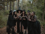 A group of six women dressed in black, wearing gothic-style clothing and hats, are standing in a forest surrounded by trees. They appear to be focused on an old book, which one of them is holding open. The mood appears mysterious and dark.