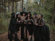 A group of six women dressed in black, wearing gothic-style clothing and hats, are standing in a forest surrounded by trees. They appear to be focused on an old book, which one of them is holding open. The mood appears mysterious and dark.