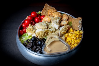 A colorful salad arranged in a bowl features cherry tomatoes, lettuce, black olives, corn, croutons, and crispy chicken pieces. A sesame-seed covered bread and a small container of dressing are also present in the bowl.