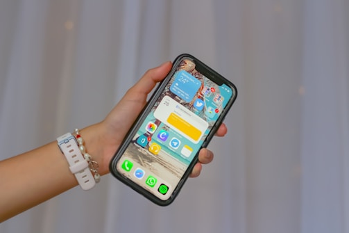 A hand holding a smartphone displaying a colorful home screen with various app icons and widgets. The phone has a black case and the person is wearing a white wristband with beaded bracelets. The background is blurred, creating a soft contrast against the focused phone.