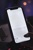 A hand is holding a smartphone with a note-taking application open on the screen. The keyboard and the text input field are visible, and it appears the user is about to type. The setting is dimly lit, possibly at night, with blurred lights in the background suggesting an indoor location.