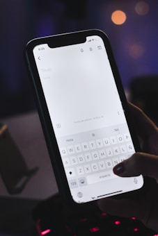 A hand is holding a smartphone with a note-taking application open on the screen. The keyboard and the text input field are visible, and it appears the user is about to type. The setting is dimly lit, possibly at night, with blurred lights in the background suggesting an indoor location.