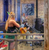 A rustic indoor scene with musicians performing. A woman with long blonde hair is playing the violin, while a man with a hat is sitting and playing the guitar, and another man is playing a string instrument at the back. The setting appears to be a small venue with brick walls, featuring a large art piece of a face on the wall.