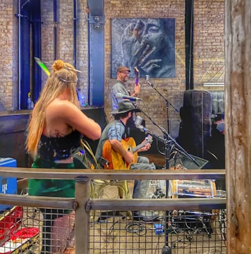 A rustic indoor scene with musicians performing. A woman with long blonde hair is playing the violin, while a man with a hat is sitting and playing the guitar, and another man is playing a string instrument at the back. The setting appears to be a small venue with brick walls, featuring a large art piece of a face on the wall.