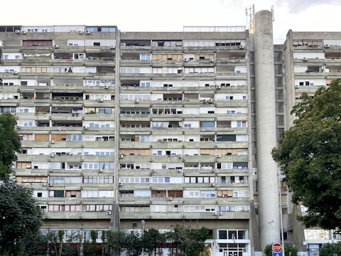 A tall, multi-story residential building with numerous balconies and air conditioning units attached to each apartment. The building's facade is a mix of concrete and glass, with varying window styles and some balconies enclosed or partially covered. Large trees with green foliage are present in the foreground, alongside a stop sign.