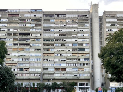 A tall, multi-story residential building with numerous balconies and air conditioning units attached to each apartment. The building's facade is a mix of concrete and glass, with varying window styles and some balconies enclosed or partially covered. Large trees with green foliage are present in the foreground, alongside a stop sign.