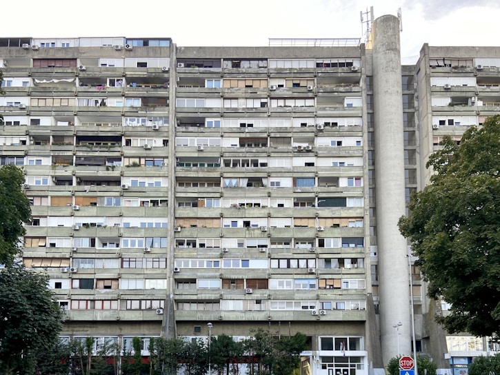 A tall, multi-story residential building with numerous balconies and air conditioning units attached to each apartment. The building's facade is a mix of concrete and glass, with varying window styles and some balconies enclosed or partially covered. Large trees with green foliage are present in the foreground, alongside a stop sign.