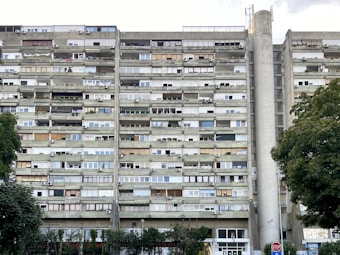A tall, multi-story residential building with numerous balconies and air conditioning units attached to each apartment. The building's facade is a mix of concrete and glass, with varying window styles and some balconies enclosed or partially covered. Large trees with green foliage are present in the foreground, alongside a stop sign.