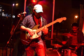 A band performs in a dimly lit venue, with the guitarist in the forefront wearing a plaid shirt and a hat, playing an electric guitar. In the background, a drummer and a bassist can be seen, all immersed in the performance. The stage is illuminated with colorful lights, creating a vibrant atmosphere.