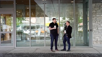 Two men are standing outside a glass entrance labeled 'Guelph City Hall,' each holding a drink. They appear to be engaged in a conversation, with one man leaning slightly and the other standing upright. The setting is urban, with reflections of trees and cars visible in the glass.