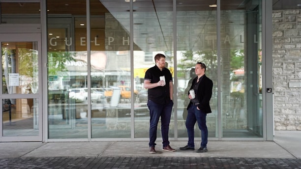 Two men are standing outside a glass entrance labeled 'Guelph City Hall,' each holding a drink. They appear to be engaged in a conversation, with one man leaning slightly and the other standing upright. The setting is urban, with reflections of trees and cars visible in the glass.