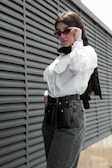 A person wearing sunglasses and a white button-up shirt stands confidently against a modern wall with horizontal ridges. They have a black scarf around their neck and a stylish belt with attachments around their jeans waistline.
