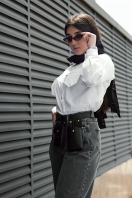 A person wearing sunglasses and a white button-up shirt stands confidently against a modern wall with horizontal ridges. They have a black scarf around their neck and a stylish belt with attachments around their jeans waistline.