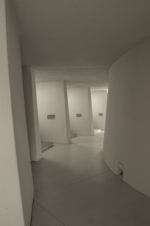 A minimalist architectural interior with smooth, curved white walls and a series of angular supports leading into a passageway. The lighting is soft and muted, creating subtle shadows and highlights on the surfaces.