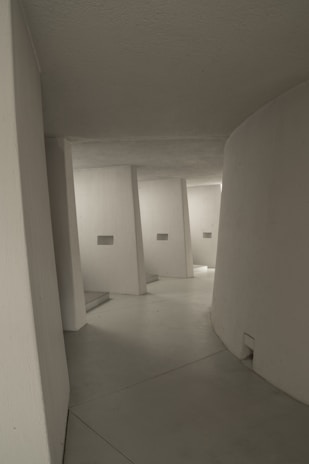 A minimalist architectural interior with smooth, curved white walls and a series of angular supports leading into a passageway. The lighting is soft and muted, creating subtle shadows and highlights on the surfaces.
