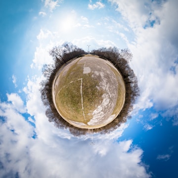A tiny planet panorama featuring a spherical view of a park-like area with barren trees circled around a concrete path. The sky is bright with scattered clouds, and the sun is shining, casting minimal shadows.